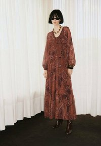 Mujer con cabello corto y oscuro que lleva gafas de sol oscuras, un largo vestido rojo con estampado paisley, collar grueso, pulseras y botas de tacón marrones, de pie en el interior.