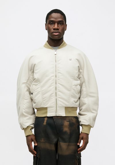 Jachetă bomber bej cu un finisaj texturat, având închidere cu fermoar, buzunare laterale și manșete și tiv din material rib. Purtați-o peste pantaloni cargo negri.