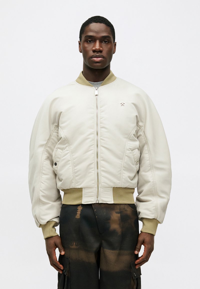 Beige bomberjack met een gestructureerde afwerking, voorzien van een ritsluiting, zijzakken en geribbelde manchetten en zoom. Gekleed over donkere cargobroeken.