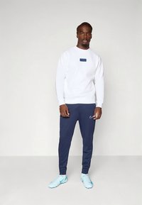 Hvit genser med blå "PARIS"-logo, kombinert med marineblå joggebukser og lyseblå treningssko. Modellen står foran en enkel bakgrunn.