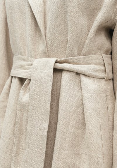 Μπεζ ρόμπα λινή με ζώνη στη μέση, με υφή ύφασμα και απλό σχέδιο, που τονίζει καθαρές γραμμές και λειτουργική κομψότητα.