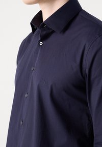 Donkerblauwe button-up shirt met een klassieke kraag, geweven stof en subtiele logodetail op de borst. Voorzien van twee knopen bij de kraag.