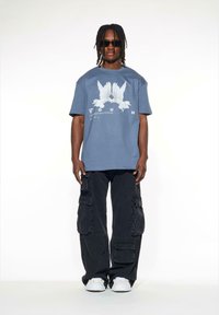 Blå bomullströja med en vit grafisk design av två fåglar, ihop med svarta cargobyxor med flera fickor. Vita sneakers används.