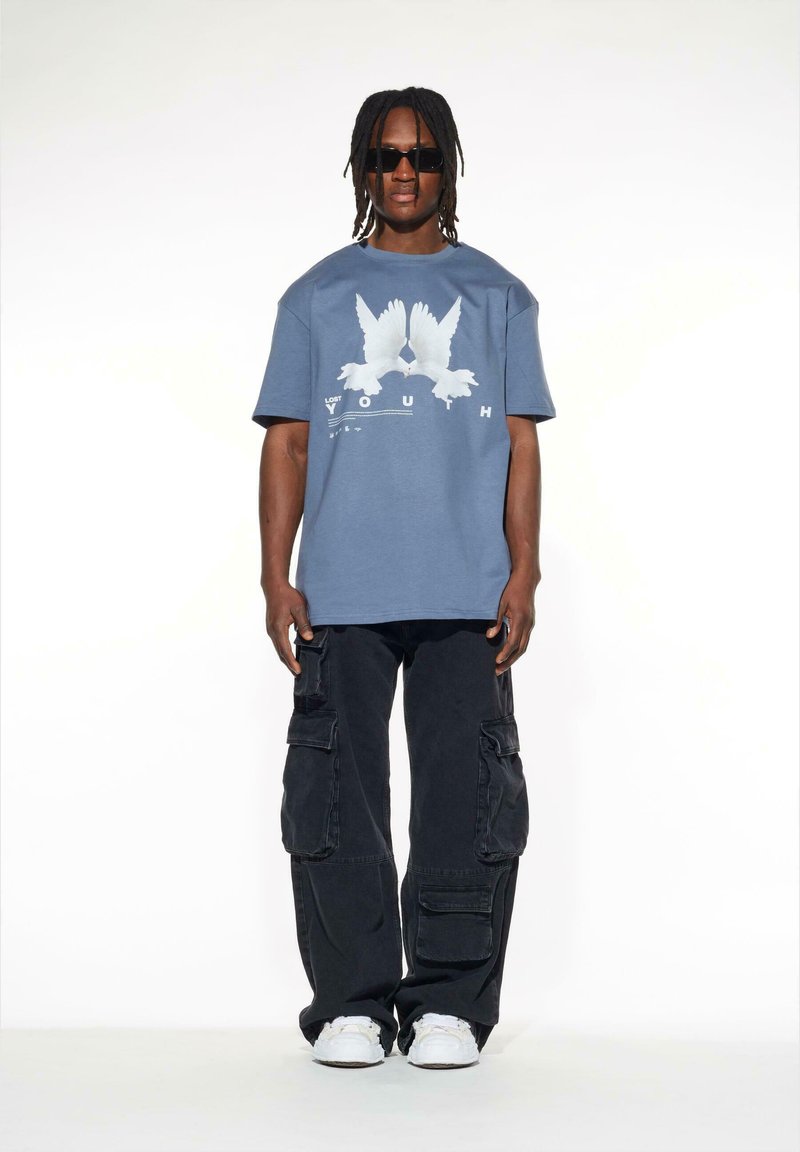 Blå bomullströja med en vit grafisk design av två fåglar, ihop med svarta cargobyxor med flera fickor. Vita sneakers används.