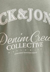 Hele roheline kangas valge tekstiga "Jack & Jones Denim Crew Collective Worldwide Established Nineteen Ninety".