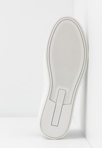 Semelle de chaussure en caoutchouc blanc avec une surface texturée, présentant une marque discrète au niveau du talon et des bords lisses, inclinée sur un fond blanc.