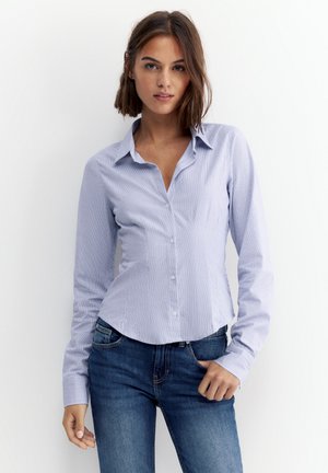 Une femme porte une chemise boutonnée rayée bleu clair et un jean bleu foncé sur un fond blanc uni.
