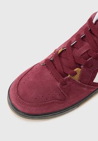 Burgundy suede sneaker with white accents, black rubber sole, and textured laces. Features perforations for breathability and a rounded toe.