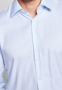 Camicia azzurra chiara con tessuto strutturato, colletto a punta e bottoni bianchi. Il design presenta una silhouette aderente.