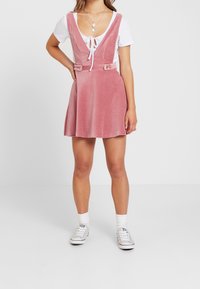 Roze ribfluwelen jurk met een diepe V-hals en verstelbare schouderbanden, voorzien van twee voorknopen en een uitlopende rok; gedragen over een wit topje.