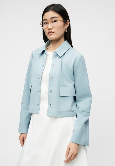 Jauna moteris dėvi šviesiai mėlyną susegamą švarką ant balto suknelės, su aiškiais akiniais rėmeliuose ir auksinėmis lankų formos auskarėmis, stovinti priešais baltą foną.