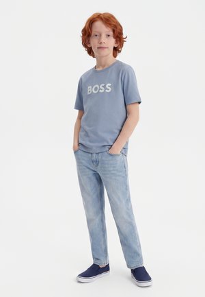 Ung rødhåret dreng iført en blå "BOSS" t-shirt, lyseblå jeans og marineblå slip-on sko, stående med hænderne i lommerne.