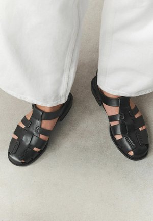 Person trägt weiße Hose und schwarze Ledersandalen und steht auf einem hellgrauen Boden.