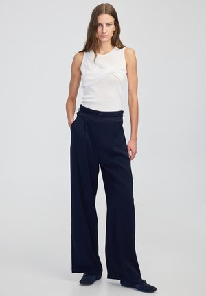 Top bianco senza maniche con un design frontale attorcigliato abbinato a pantaloni ampi in vita alta di colore blu navy. Tessuto liscio, pieghe sottili in vita.