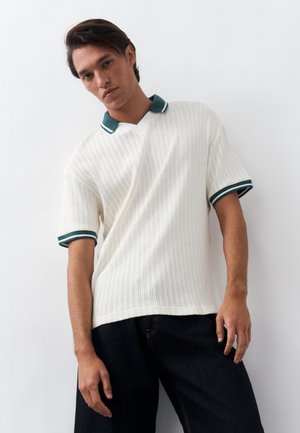 Jaunas vyras dėvi baltą trumpomis rankovėmis megztą polo marškinėlį su tamsiai žalia apykakle ir rankovių apvadais, stovintis priešais blankią baltą foną.