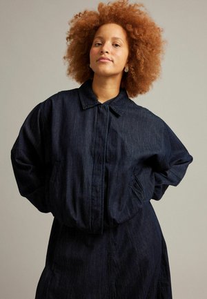 Kvinde med krøllet rødt hår iført en mørkeblå oversized denimjakke med hænderne i lommerne mod en ensfarvet beige baggrund.