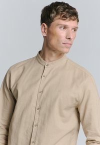 Beige, langetermijnsleeve shirt met een bandkraag en vijf knopen, met een subtiele geborduurde logo op de borst. Glad textuur.