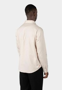 Lys beige langermet skjorte med krage, en knapp ved mansjettene, og buet søm. Teksturert stoff med en subtil glans.