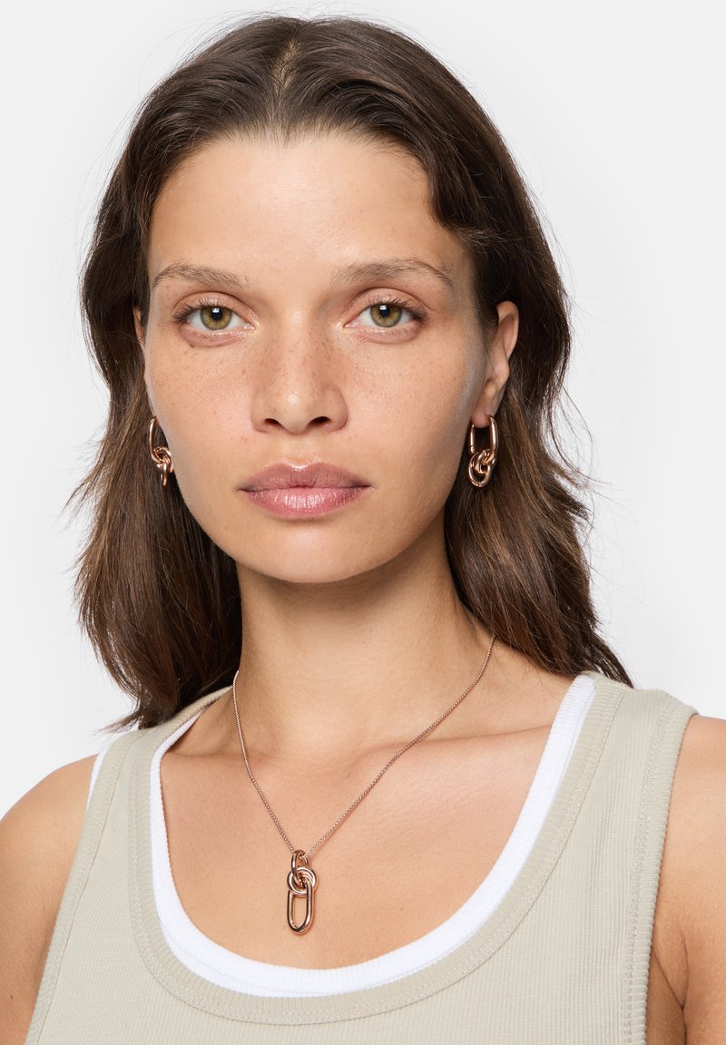 Halskette mit Anhänger, die aus miteinander verbundenen Metallelementen in Roségold besteht, getragen über einem hellbeigen Tanktop mit einem weißen Halsausschnitt.