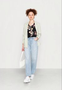 Blazer verde claro sobre uma blusa preta com flores, combinado com jeans claros de cintura alta e ténis brancos, segurando uma bolsa branca.