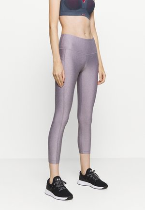 Femme portant un soutien-gorge de sport sombre, des leggings courts taille haute violets et des chaussures de sport noires, debout sur un fond blanc.