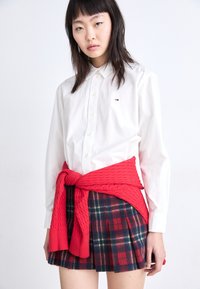 White button-up shirt with a logo on the chest, paired with a red knitted sweater tied around the waist and a plaid pleated skirt.