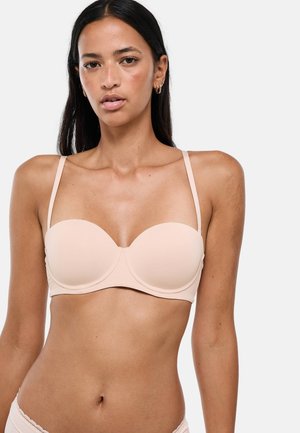 Femme aux longs cheveux foncés portant un soutien-gorge beige clair sans bretelles avec des bretelles fines, debout devant un fond clair uni.