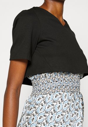 Frau trägt ein schwarzes, kurzes, bauchfreies Oberteil über einem weißen Kleid mit blauem und schwarzem Blumenmuster und geraffter Taille.