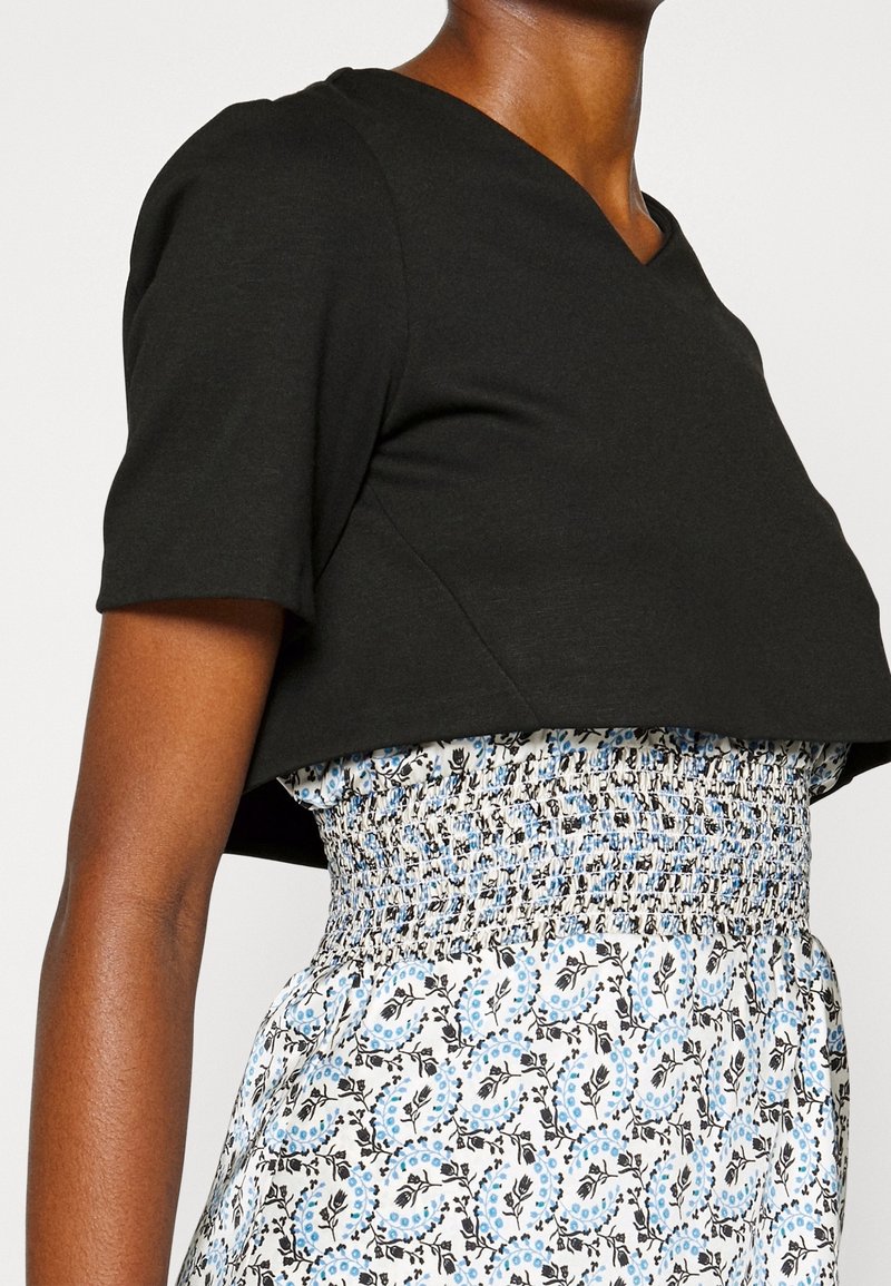 Femme portant un haut court noir à manches courtes sur une robe blanche avec un motif floral bleu et noir et une taille smockée.