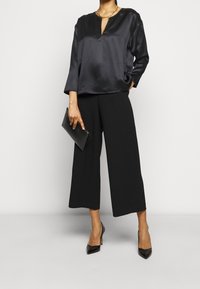 Blouse en satin noir à encolure à trou de serrure, associée à un pantalon large noir. La tenue est accessoirisée d'une pochette noire et de talons hauts.