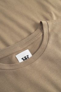 Beige Rundhals-T-Shirt mit sichtbaren Rippkragen-Nähten und einem weißen Etikett mit einem auffälligen schwarzen Logo im Nacken.