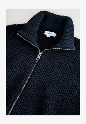 Jūras flotes zip-up džemperis ar teksturētu adījuma dizainu, ribinātu apkakli un sudraba rāvējslēdzēju. Marķējums redzams apkakles iekšpusē.