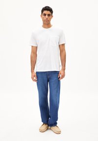 Weißes Baumwoll-T-Shirt mit einer Brusttasche, kombiniert mit blauen Jeans. Lockere Passform, minimalistisches Design und beigefarbene Slipper sorgen für Komfort.