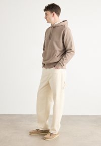 Bruine hoodie met een voorzak, gecombineerd met beige broek met wijde pijpen en lichtbruine instappers. Eenvoudig ontwerp, zachte textuur.
