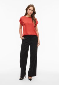 Rød silkebluse med rullede ærmer, parret med sorte vidde bukser og sorte højhælede sko med guld kæde detaljer.