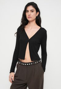 Μαύρη, υφή με μακριά μανίκια ζακέτα με ντεκολτέ σε σχήμα V και κλείσιμο με κουμπιά. Κοντή εφαρμογή με κυματιστό τελείωμα. Φορεμένη πάνω από ψηλόμεσα παντελόνια με ζώνη με τρουκς.