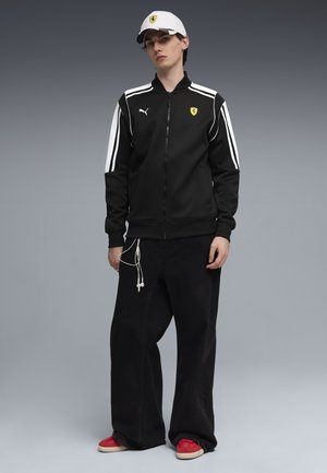 Young man stands wearing black Puma and Ferrari zip-up jacket, black wide-leg pants, white Ferrari cap, and red sneakers against gray background.
