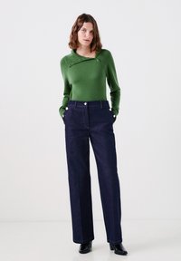 Maglione verde lavorato a maglia con un colletto diagonale con bottoni abbinato a pantaloni in denim a gamba larga blu scuro con dettagli in bottone metallico.
