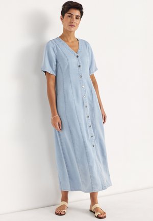 Frau trägt ein hellblaues Jeanskleid mit Knopfleiste vorne, kurzen Ärmeln und beigen Sandalen, steht vor einer weißen Wand.