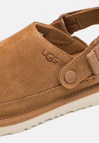 Bruna mockasiner i mocka med en sömmad beige sula, utrustad med ett elastiskt band, UGG-logga och en texturerad detalj på sidan.