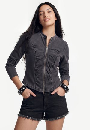 Giovane donna con capelli lunghi e scuri, indossa una giacca grigia aderente con zip, pantaloncini di jeans neri sfrangiati e bracciali in pelle borchiati.