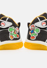 Zapatillas negras con suelas amarillas, que presentan parches de personajes de plástico de Yoshi, Mario y Toad. El diseño incluye acentos blancos y material de malla.