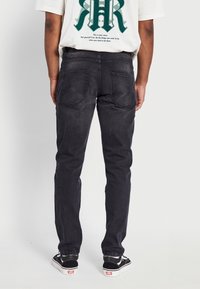 Una persona sta in piedi voltata di spalle, indossando jeans neri, sneakers bianche e nere, e una maglietta bianca con testo verde e nero sulla schiena.