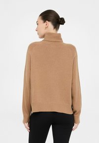 Donna con capelli castani raccolti in un bun, che indossa orecchini dorati, un maglione a collo alto color cammello e pantaloni neri, rivolta a sinistra su uno sfondo bianco.