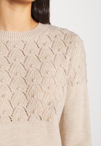 Beige gebreide trui met een gestructureerd ontwerp met opengewerkte patronen en kleine bobbels. Ronde halslijn en lange mouwen.