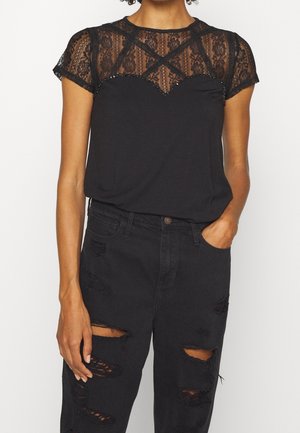 Vrouw draagt zwart kanten korte mouwen top met kralen versiering bij de hals en zwart gescheurde high-waist jeans, één hand in de zak tegen een witte achtergrond.
