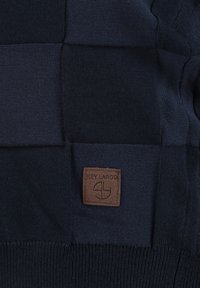 Granatowy, dzianinowy materiał w patchworkowym stylu z małą, brązową skórzaną metką wytłoczoną napisem "KEY LARGO" oraz okrągłym logo.