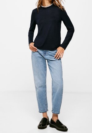 Femme portant un haut noir à manches longues, un jean bleu clair retroussé et des mocassins noirs, debout les mains sur les hanches devant un fond uni.
