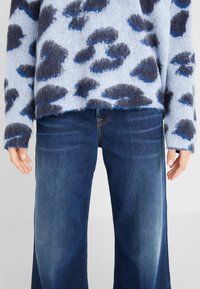 Personne portant un pull duveteux bleu à motif léopard et un jean bleu foncé à jambes larges, les mains détendues le long du corps.