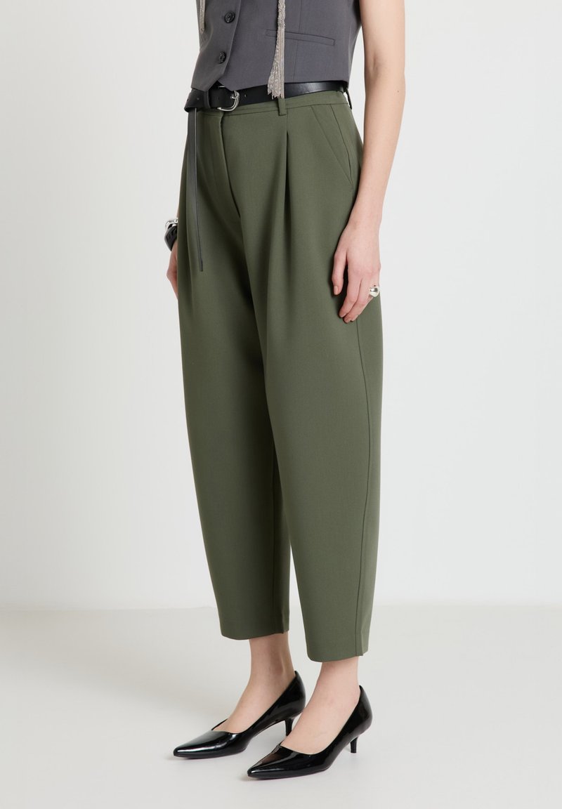 Donna che indossa pantaloni a vita alta color oliva con gamba affusolata, cintura nera, top grigio senza maniche e scarpe nere con tacco basso e punta, in piedi su pavimento bianco.